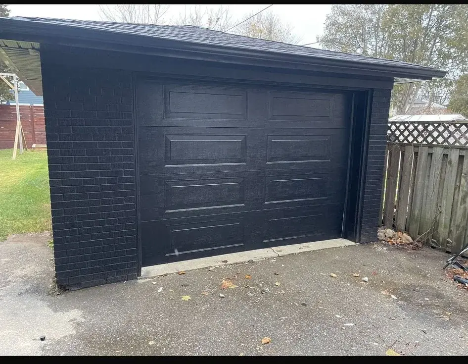 Black raised-panel garage door installed on a detached garage with dark masonry