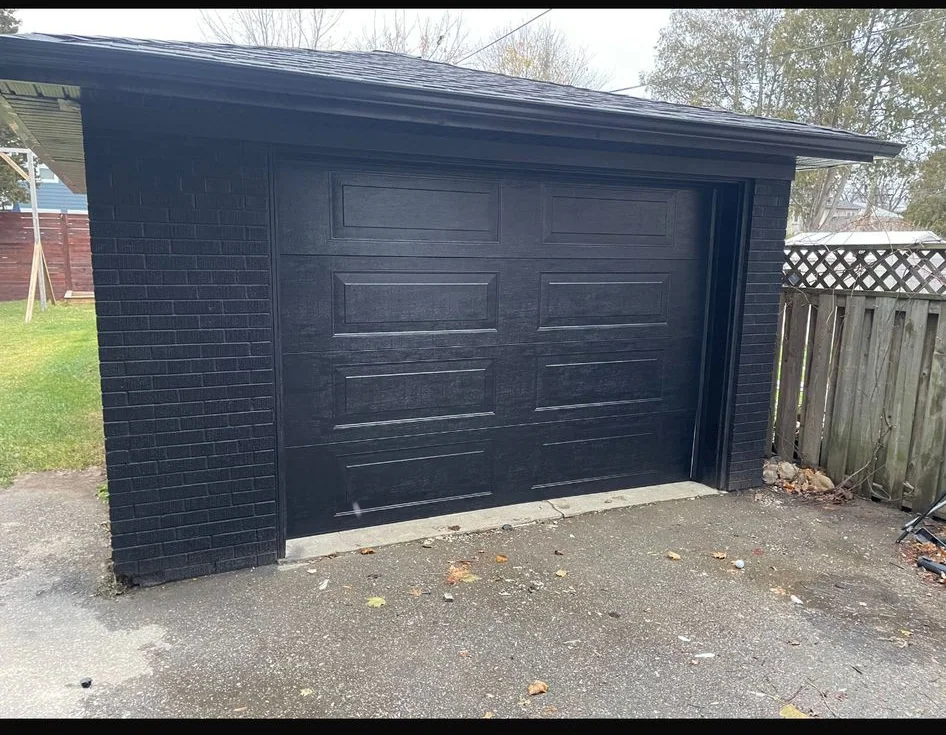 Black raised-panel garage door installed on a detached garage with dark masonry